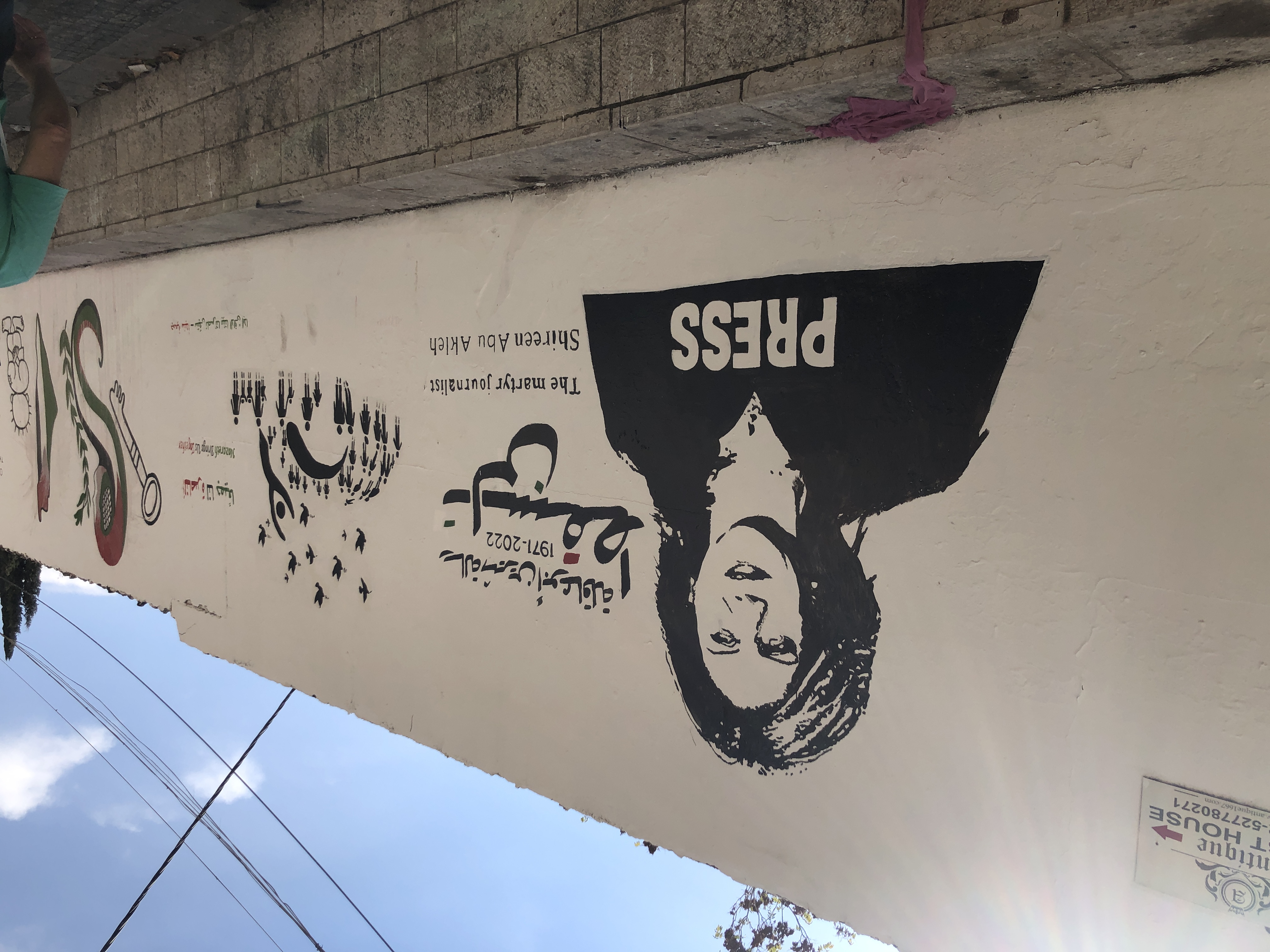 Čiernobiele grafiti ženy vo veste s nápisom PRESS a arabským textom na bielom múre v Nazarete