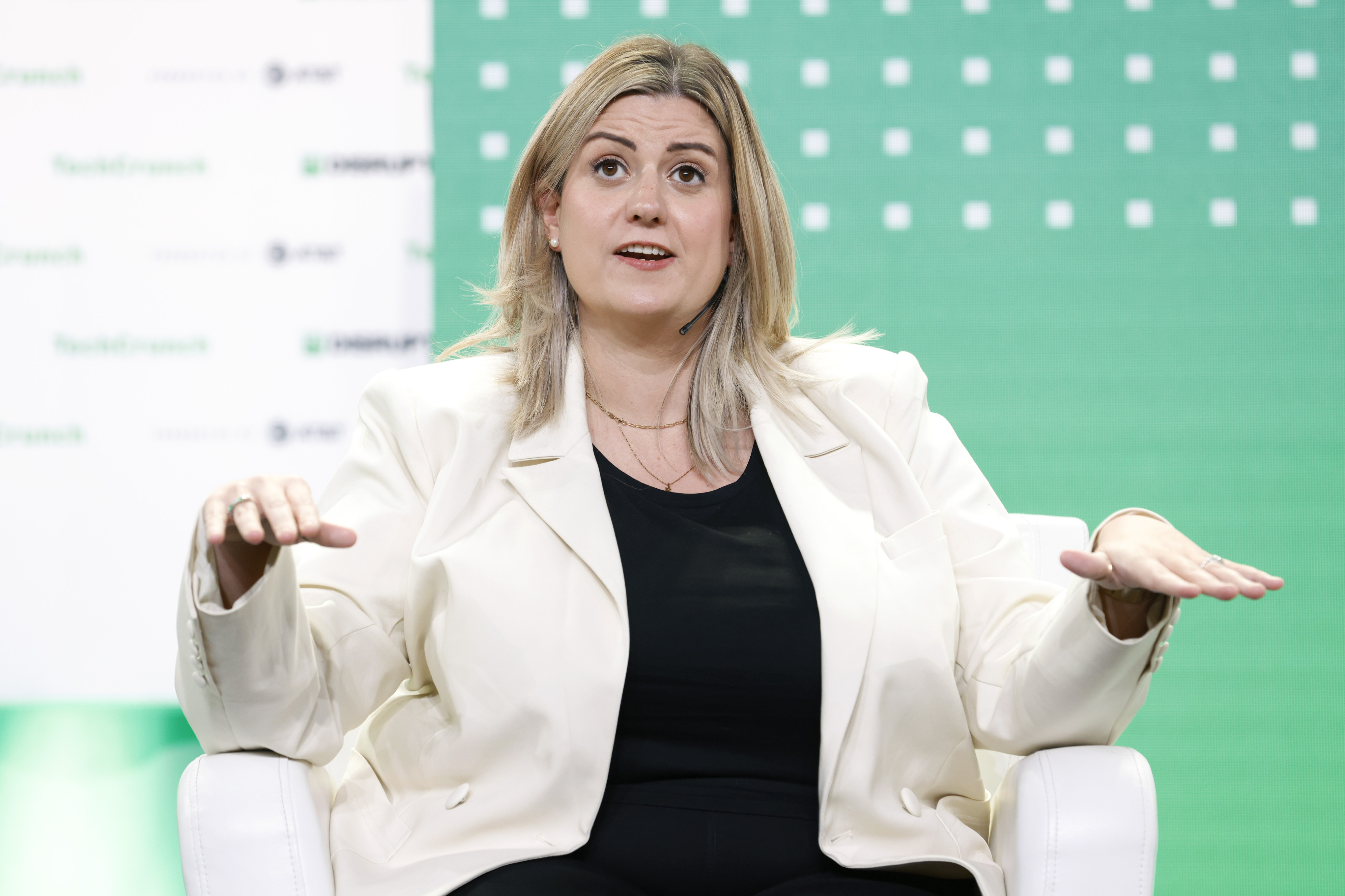 Žena v bielom saku sedí na pódiu konferencie pred zeleným panelom a gestikuluje rukami